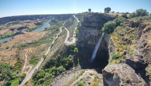 A panoramic view of a canyon with a waterfall, winding road, and lush greenery along the riverbank.