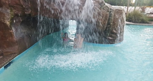 A person emerges from a waterfall in a pool, with water splashing around them and a rocky structure in the background.