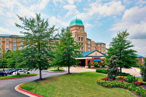 A hotel with a green dome, surrounded by trees and colorful flowers, under a partly cloudy sky.