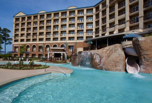 A resort pool area with a water slide, surrounded by landscaped gardens and a multi-story building in the background.