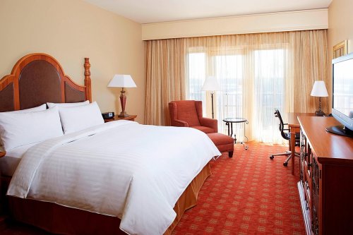 A cozy hotel room featuring a king bed, armchair, desk, and large windows with sheer curtains.