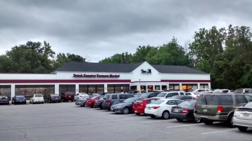 Exterior of Dutch Country Farmers Market with a parking lot filled with cars under a cloudy sky.