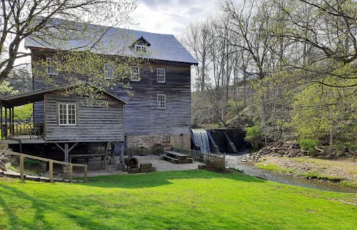A rustic wooden mill beside a stream, surrounded by greenery and trees under a clear sky.