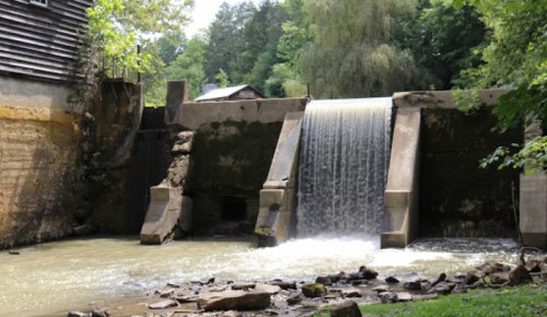 A small waterfall flows over a concrete dam, surrounded by lush greenery and a rustic wooden structure nearby.