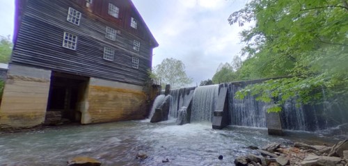 A historic wooden mill beside a flowing river and a small waterfall, surrounded by lush greenery.