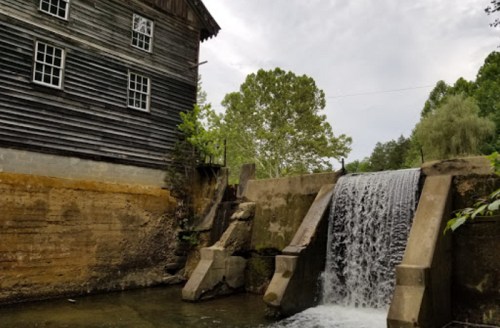 A historic wooden mill beside a flowing waterfall, surrounded by lush greenery and a cloudy sky.