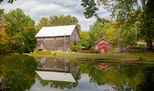 A rustic wooden mill and a small shed with a red wheel, reflected in a calm pond surrounded by trees.