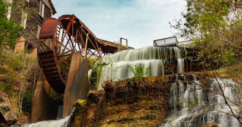 A scenic view of a watermill with a large wheel, cascading waterfalls, and lush greenery surrounding it.