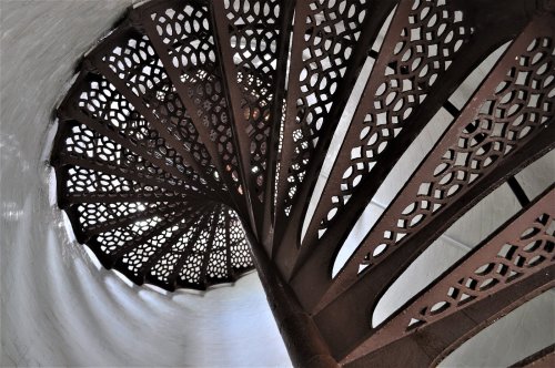 A spiral staircase with intricate metalwork, viewed from above, leading down into a light-filled space.