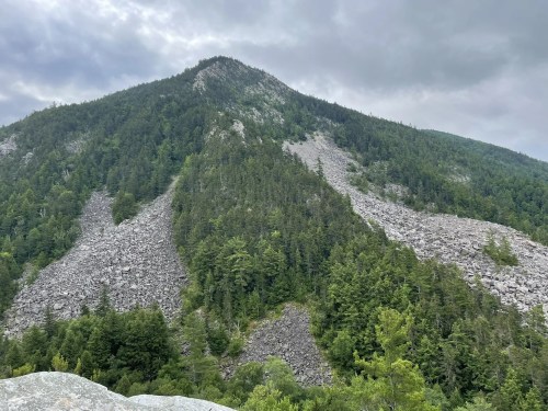 A rocky mountain slope covered in trees under a cloudy sky, showcasing a mix of greenery and rocky terrain.