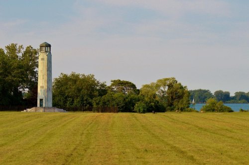 A tall, white lighthouse stands near a grassy field, surrounded by trees and overlooking a calm body of water.