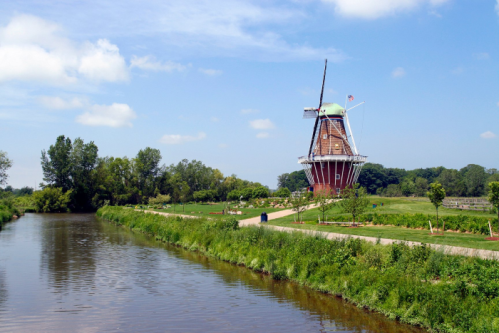 A scenic view of a windmill beside a calm river, surrounded by lush greenery and a clear blue sky.