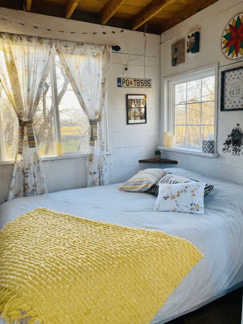 Cozy bedroom with a large bed, yellow blanket, floral pillows, and bright windows showcasing a scenic view.