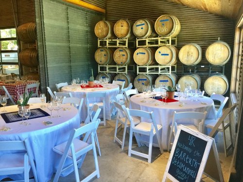 A rustic dining area with round tables, wine barrels in the background, and a welcome sign for an event.