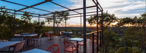 A scenic outdoor dining area with tables and chairs, overlooking a sunset and lush landscape.