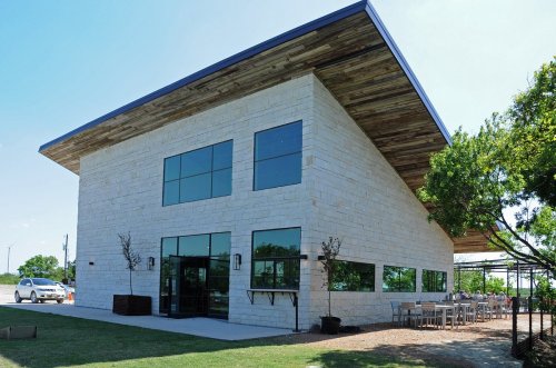 Modern building with a stone exterior, large windows, and a wooden roof, surrounded by greenery and outdoor seating.