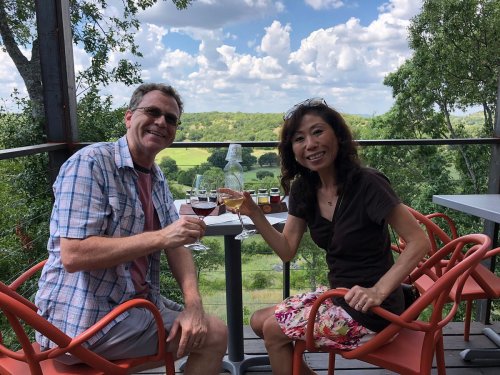 A man and woman toast with drinks on a balcony overlooking a scenic landscape with trees and clouds.