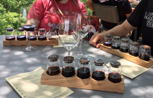 A table with wine tasting flights in glass jars, accompanied by empty wine glasses and menus, set outdoors.