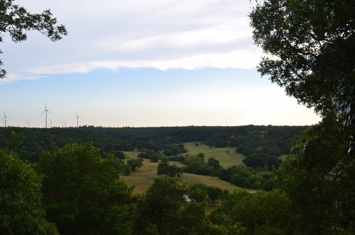 A scenic view of rolling green hills and trees, with wind turbines visible in the distance under a cloudy sky.