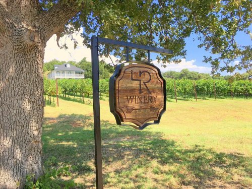 A wooden sign for "LR Winery" hangs from a post near a vineyard, with a house visible in the background under a blue sky.