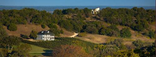 A scenic view of rolling hills with a vineyard, a house, and a modern building in the distance under a clear blue sky.