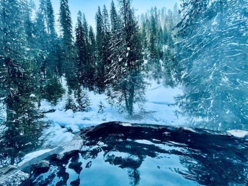 A steaming hot spring surrounded by snow-covered trees and a winter landscape.