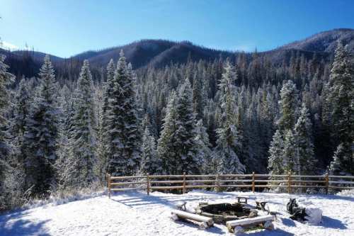 Snow-covered trees surround a wooden fence and fire pit, with mountains in the background under a clear blue sky.