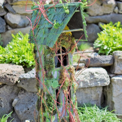 A whimsical birdhouse covered in moss and vines, nestled among stones and greenery in a garden setting.