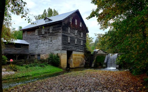A rustic wooden mill beside a small waterfall, surrounded by trees and autumn leaves.