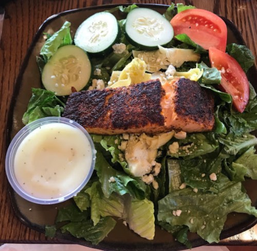A plate with a seasoned salmon fillet on a bed of mixed greens, cucumbers, tomatoes, and a small cup of dressing.