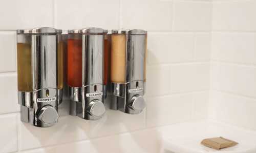 Three chrome shower dispensers mounted on a tiled wall, labeled for shower gel, shampoo, and conditioner.