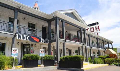 A building with a sign reading "Wine Zone," featuring balconies and a welcoming entrance surrounded by greenery.