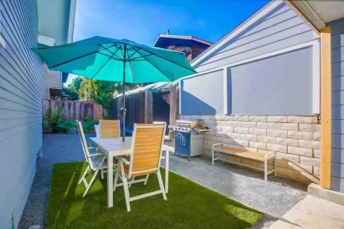 A cozy outdoor patio with a table, chairs, and a large umbrella, surrounded by greenery and a grill nearby.