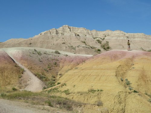 A person stands on colorful, layered hills under a clear blue sky in a scenic landscape.