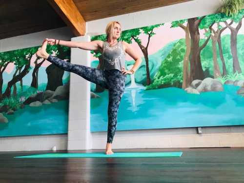 A person practicing yoga in a studio, balancing on one leg with a scenic mural of trees and water in the background.