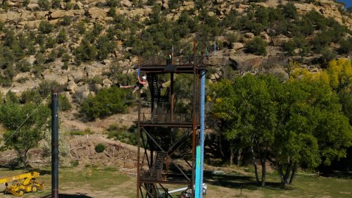 A person zip-lining from a tall metal tower, surrounded by rocky terrain and greenery.