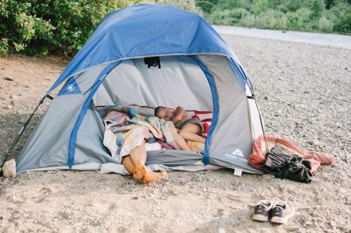 Two people sleeping inside a blue tent on a gravel beach, covered with colorful blankets. Shoes are nearby.