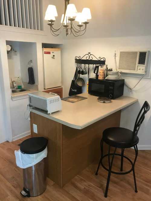 A small kitchen with a countertop, bar stool, microwave, and a chandelier above, featuring a trash can and air conditioner.