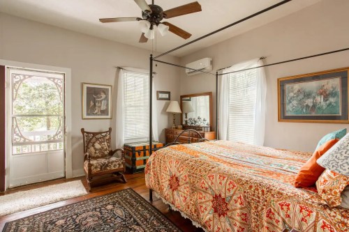 Cozy bedroom with a four-poster bed, colorful quilt, armchair, and natural light from windows and door.