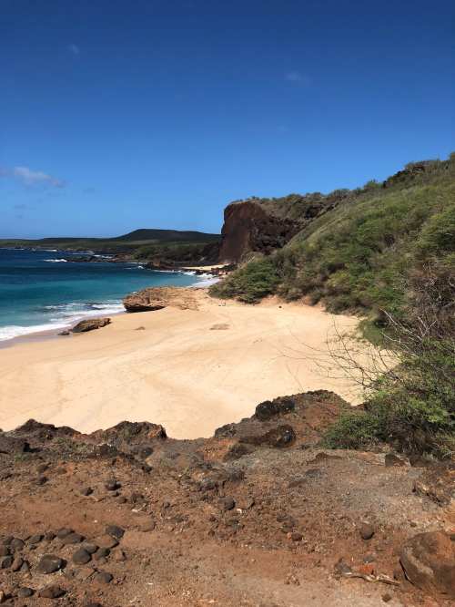 Scenic beach view with golden sand, rocky cliffs, and clear blue water under a bright blue sky.
