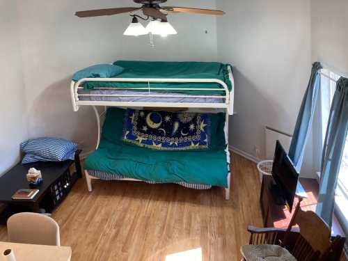 A bright room with a bunk bed, a green blanket, a table, and a chair, featuring wooden flooring and large windows.