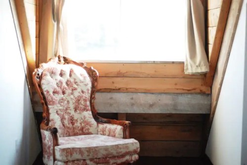 A vintage upholstered chair sits in a cozy, sunlit corner with wooden walls and curtains.