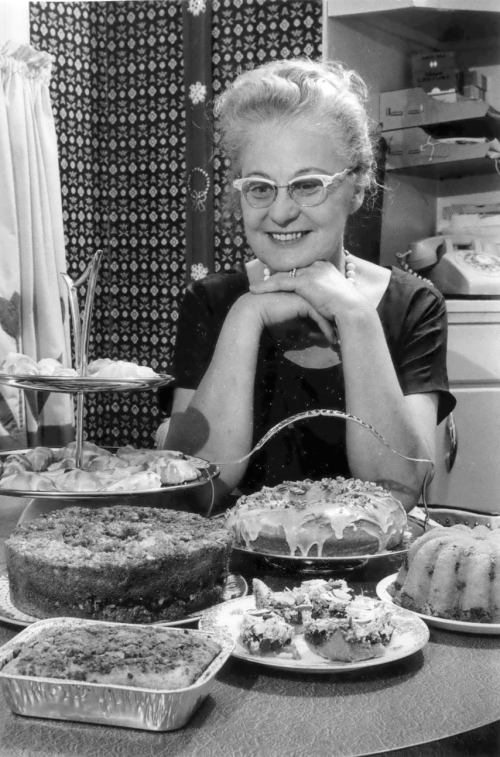 A smiling woman with glasses poses at a table filled with various baked goods and desserts in a cozy kitchen setting.