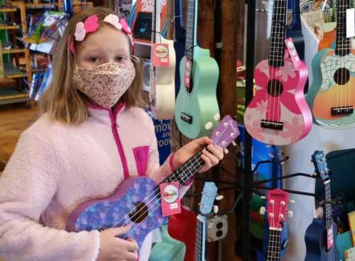 A girl in a pink jacket and mask holds a purple ukulele in a store filled with colorful ukuleles.