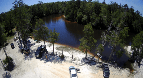 Aerial view of a serene pond surrounded by trees, with parked cars along the sandy shore.