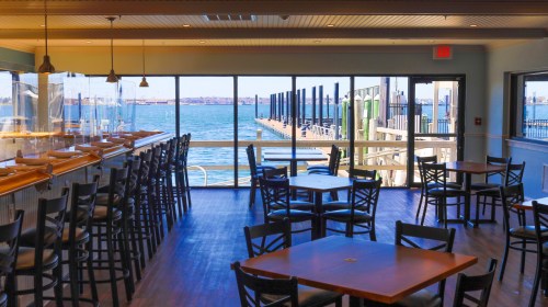 Bright restaurant interior with wooden tables and chairs, overlooking a waterfront dock and blue water through large windows.
