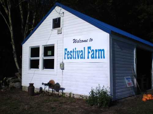 A white building with a blue roof and "Welcome to Festival Farm" sign on the side, surrounded by greenery.