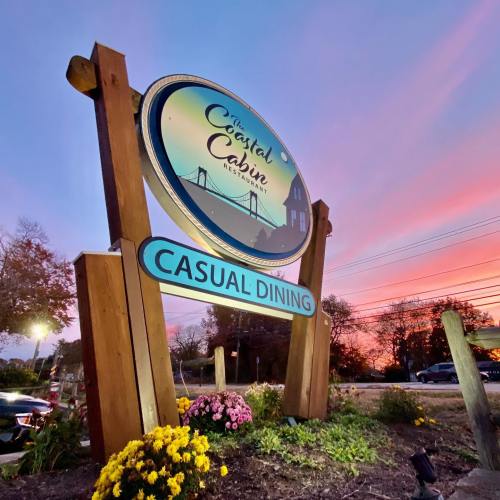 Sign for "The Coastal Cabin" restaurant at sunset, featuring a logo and "Casual Dining" in blue lettering.