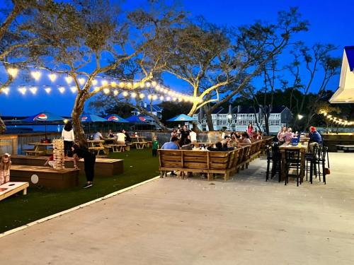 Outdoor dining area at dusk, featuring string lights, wooden seating, and patrons enjoying the atmosphere by the water.