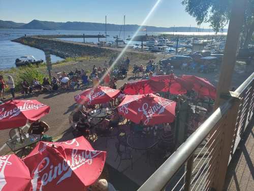 A waterfront scene with people gathered under red umbrellas, enjoying a sunny day by the marina with boats in the background.